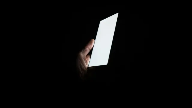 Hand hält ein hell leuchtendes Smartphone in völliger Dunkelheit – Poppen.de oder Joyclub im Vergleich